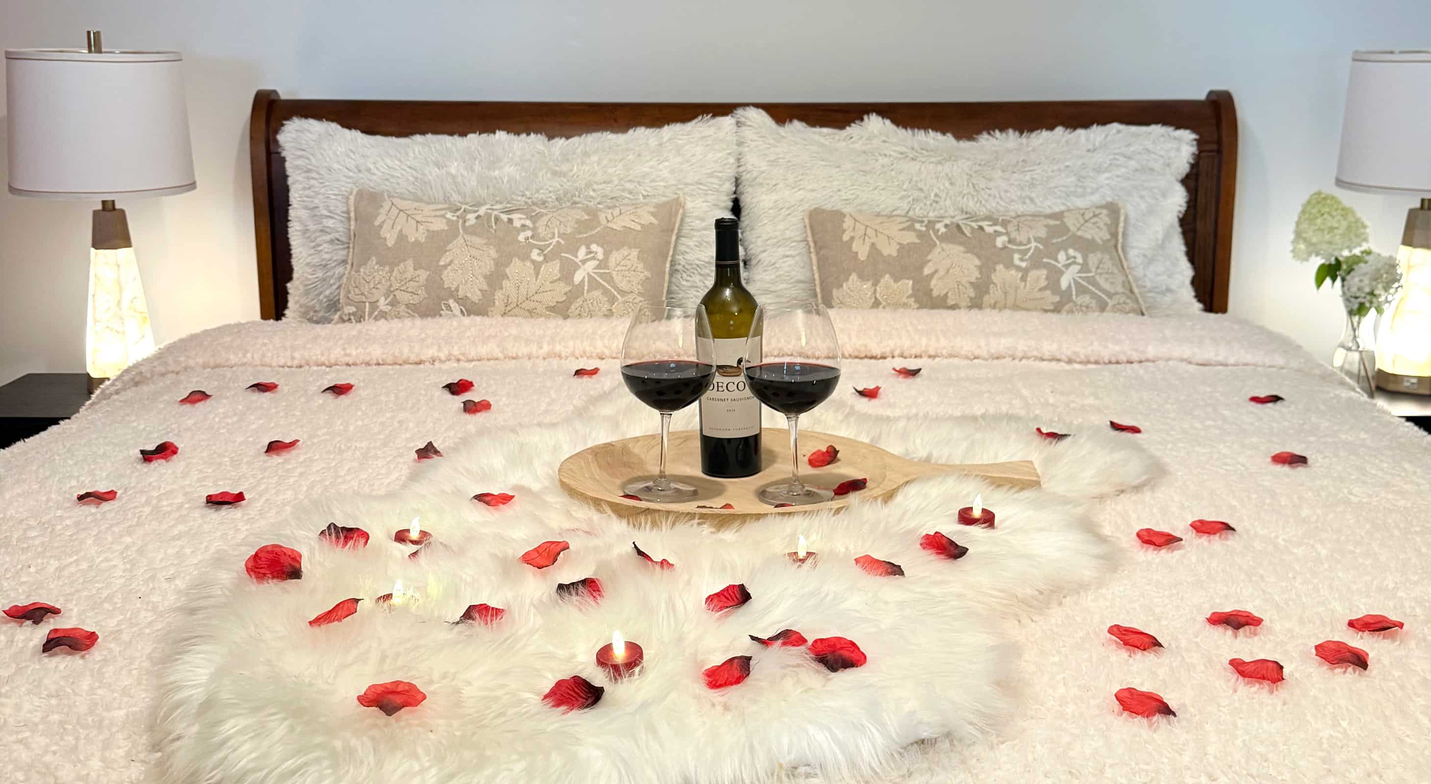 Two wine glasses, bottle of wine, rose petals, and tea lights on a bed